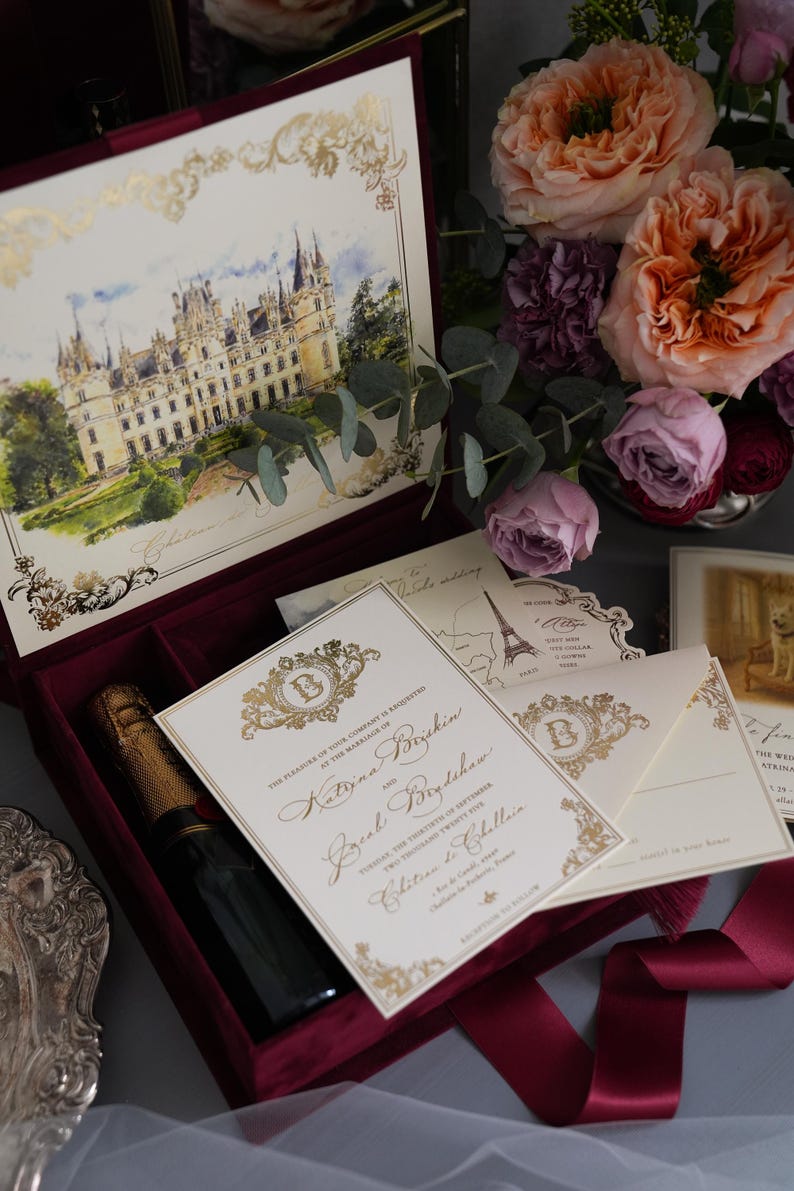 K&ouml;nnte beinhalten: Hochzeitseinladungs-Set mit Schlossillustration, Goldakzenten und einer bordeauxroten Samtbox. Enth&auml;lt Einladungskarten, Umschl&auml;ge und eine Champagnerflasche. Blumen im Hintergrund.