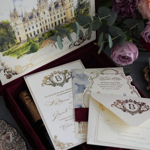 K&ouml;nnte beinhalten: Eine bordeauxfarbene Samtbox mit Hochzeitseinladungen, einer Champagnerflasche und Schreibwaren. Die Einladungen zeigen goldene Schrift und eine Schlossillustration. Blumenschmuck mit rosa und lila Rosen erg&auml;nzt die Szene.