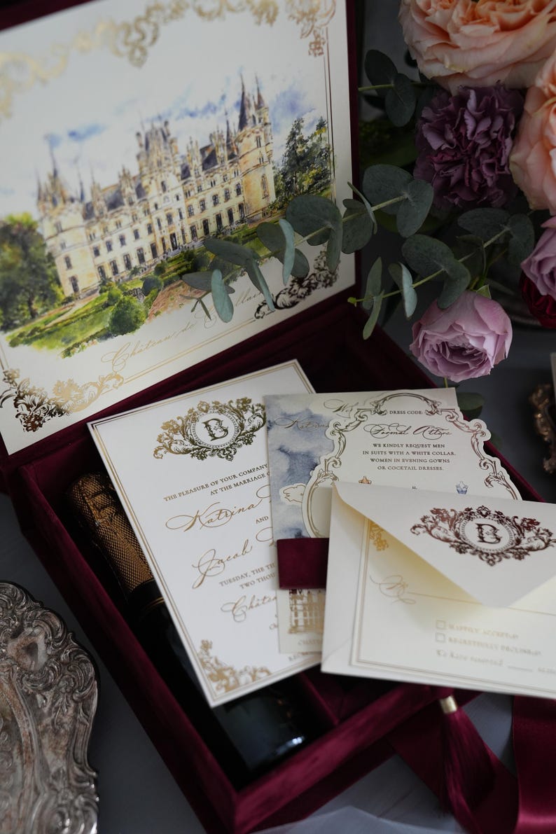 K&ouml;nnte beinhalten: Eine bordeauxfarbene Samtbox mit Hochzeitseinladungen, einer Champagnerflasche und Schreibwaren. Die Einladungen zeigen goldene Schrift und eine Schlossillustration. Blumenschmuck mit rosa und lila Rosen erg&auml;nzt die Szene.