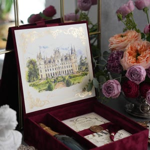 K&ouml;nnte beinhalten: Eine bordeauxfarbene Samt-Pr&auml;sentationsbox mit einem goldgerahmten Aquarelldruck eines Schlosses. Im Inneren sind eine Champagnerflasche und Einladungskarten zu sehen. Die Box ist ge&ouml;ffnet und zeigt ihren Inhalt, mit einem Blumenstrau&szlig; im Hintergrund.