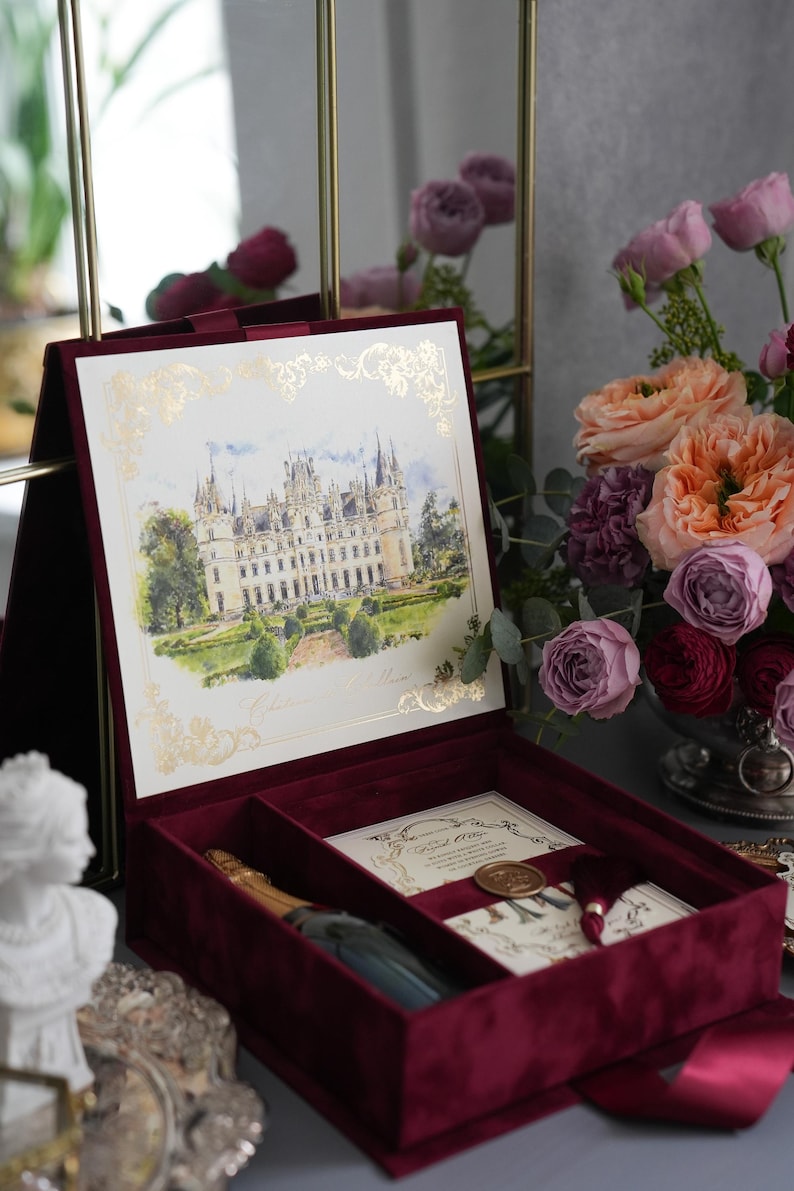 K&ouml;nnte beinhalten: Eine bordeauxfarbene Samt-Pr&auml;sentationsbox mit einem goldgerahmten Aquarelldruck eines Schlosses. Im Inneren sind eine Champagnerflasche und Einladungskarten zu sehen. Die Box ist ge&ouml;ffnet und zeigt ihren Inhalt, mit einem Blumenstrau&szlig; im Hintergrund.