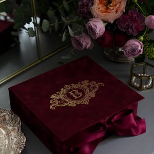K&ouml;nnte beinhalten: Eine weinrote Samtbox mit goldenem, verziertem Monogramm und passendem Band. Die Box wird mit einem Strau&szlig; Rosen in Rosa-, Pfirsich- und Burgundert&ouml;nen und einem dekorativen Spiegel pr&auml;sentiert.