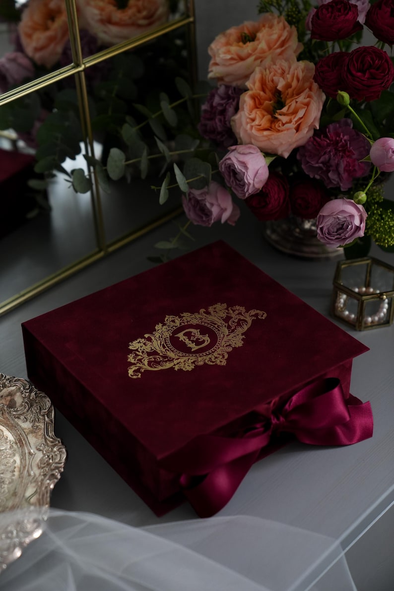 K&ouml;nnte beinhalten: Eine weinrote Samtbox mit goldenem, verziertem Monogramm und passendem Band. Die Box wird mit einem Strau&szlig; Rosen in Rosa-, Pfirsich- und Burgundert&ouml;nen und einem dekorativen Spiegel pr&auml;sentiert.