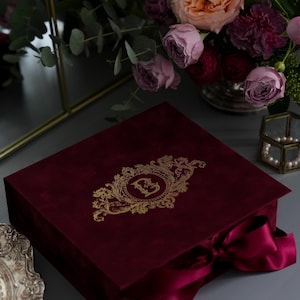 K&ouml;nnte beinhalten: Eine weinrote Samtbox mit einem goldenen, verzierten Design und einem passenden Band. Die Box ist geschlossen und steht auf einer grauen Oberfl&auml;che, mit einem Blumenstrau&szlig; im Hintergrund. Das Design auf der Box scheint ein Monogramm zu sein.