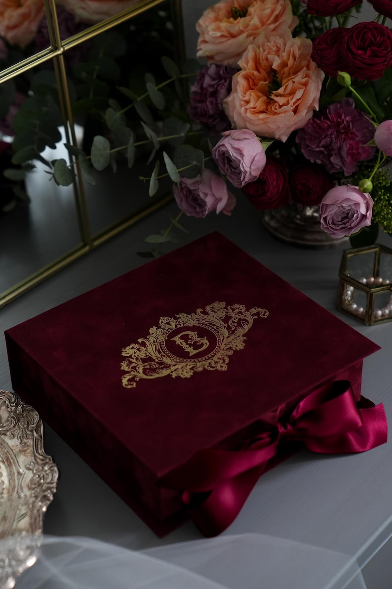 K&ouml;nnte beinhalten: Eine weinrote Samtbox mit einem goldenen, verzierten Design und einem passenden Band. Die Box ist geschlossen und steht auf einer grauen Oberfl&auml;che, mit einem Blumenstrau&szlig; im Hintergrund. Das Design auf der Box scheint ein Monogramm zu sein.