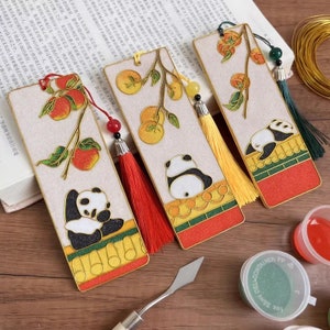 Op de afbeelding: Drie handgemaakte bladwijzers met een gouden rand en een panda-ontwerp. Elke bladwijzer heeft een tak met rode en oranje vruchten en een kwast.