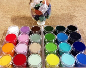 May include: A clear plastic container filled with colorful gemstones, sitting on a table with 24 small jars of colorful sand. The jars are arranged in two rows of 12, with each jar containing a different color of sand.