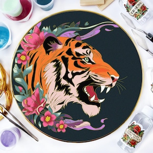 Puede incluir: Un bastidor de bordado circular con una ilustración detallada de una cabeza de tigre de perfil con la boca abierta, rodeada de flores rosas y hojas verdes sobre un fondo oscuro, junto a suministros de arte y un frasco etiquetado ERSENN.