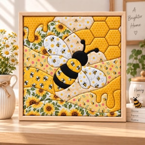 Op de afbeelding: Een vierkant ingelijst wandkunstwerk met een centrale zwart-gele bij omringd door een patchworkontwerp van zonnebloemen, kleine bijtjes en gele honingraatpatronen. Het kunstwerk gebruikt een levendig kleurenpalet van geel, wit en zwart.