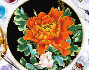 Puede incluir: Un plato decorativo redondo negro con borde dorado, con un gran diseño floral de peonía naranja y amarilla. La flor está rodeada de hojas verdes y azules, y una flor rosa más pequeña. El plato está sobre una superficie blanca.