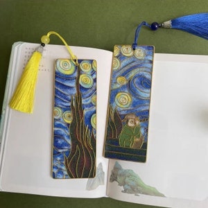 Puede incluir: Dos marcapáginas de color dorado con un diseño de noche estrellada. El marcapáginas de la izquierda tiene un cielo nocturno estrellado azul y amarillo con un árbol verde oscuro en primer plano. El marcapáginas de la derecha tiene un cielo nocturno estrellado azul y amarillo con una figura verde en primer plano. Ambos marcapáginas tienen un borla.