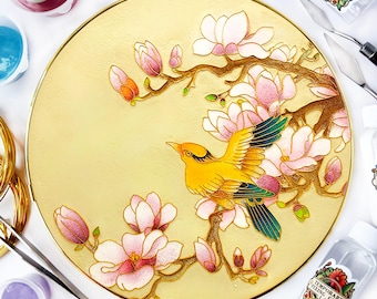 Può includere: Un piatto rotondo con bordo dorato con un disegno dipinto di un uccello giallo appollaiato su un ramo con fiori di magnolia rosa. Il piatto è circondato da vari materiali artistici, tra cui bottiglie di vernice, un coltello da tavolozza e una bottiglia di colla di fissaggio temporanea.