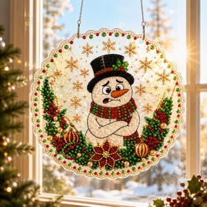 Pode incluir: Um apanhador de sol circular com um design de pintura de diamante de um boneco de neve com cartola preta e cachecol listrado de vermelho e verde, rodeado por flocos de neve dourados, folhas de azevinho, bagas vermelhas e ornamentos dourados.