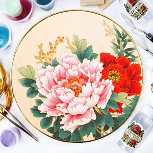 Pode incluir: Um prato decorativo redondo, creme, com uma borda dourada, apresentando um design floral detalhado. O design inclui uma grande peônia rosa, uma flor vermelha e folhas verdes. Materiais de artesanato, incluindo cola e ferramentas, circundam o prato.