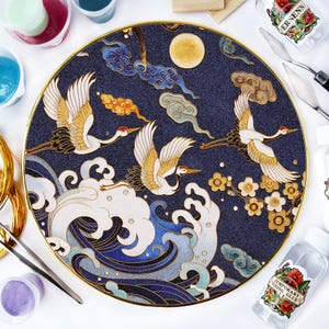 May include: A circular navy blue decorative plate with a gold edge. The plate showcases a detailed illustration of cranes flying over stylized waves and clouds. The design incorporates gold, white, and blue, with a full moon in the background.