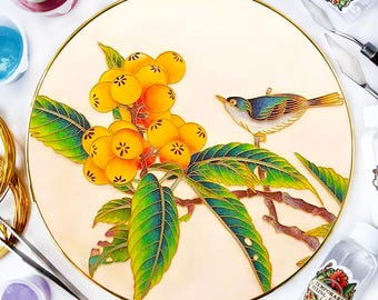 Può includere: Un piatto rotondo bianco con un bordo dorato presenta un disegno dipinto a mano di un uccello appollaiato su un ramo con foglie verdi e frutti gialli. Il piatto è circondato da vari materiali artistici, tra cui bottiglie di vernice, pennelli e una tavolozza.