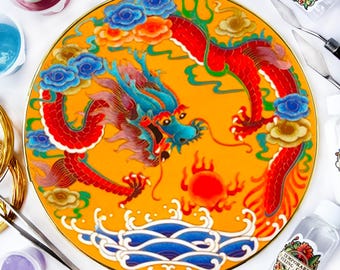May include: A round, yellow plate with a gold rim features a colorful dragon design. The dragon is red, blue, and green with a yellow background. The plate is surrounded by various craft supplies, including paint bottles, brushes, and a spatula.
