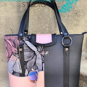 Puede incluir: Bolso tote negro con detalles de tela con estampado floral rosa. El bolso tiene dos asas negras y una cremallera. Las gafas de sol est&aacute;n guardadas en el bolsillo lateral.