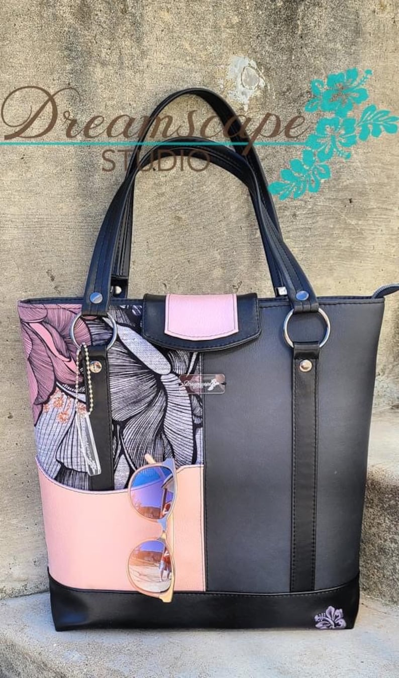 Puede incluir: Bolso tote negro con detalles de tela con estampado floral rosa. El bolso tiene dos asas negras y una cremallera. Las gafas de sol est&aacute;n guardadas en el bolsillo lateral.