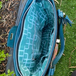 Puede incluir: Un bolso tote azul turquesa con una cremallera negra y un forro estampado en blanco y negro. El bolso tiene un asa superior y una correa para el hombro.