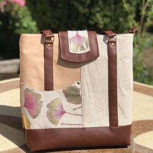 Puede incluir: Un bolso tote de cuero marr&oacute;n con un panel de tela beige y blanco con un dise&ntilde;o de hoja de ginkgo. El bolso tiene dos correas de cuero marr&oacute;n y una solapa de cuero marr&oacute;n con un dise&ntilde;o de hoja de ginkgo.