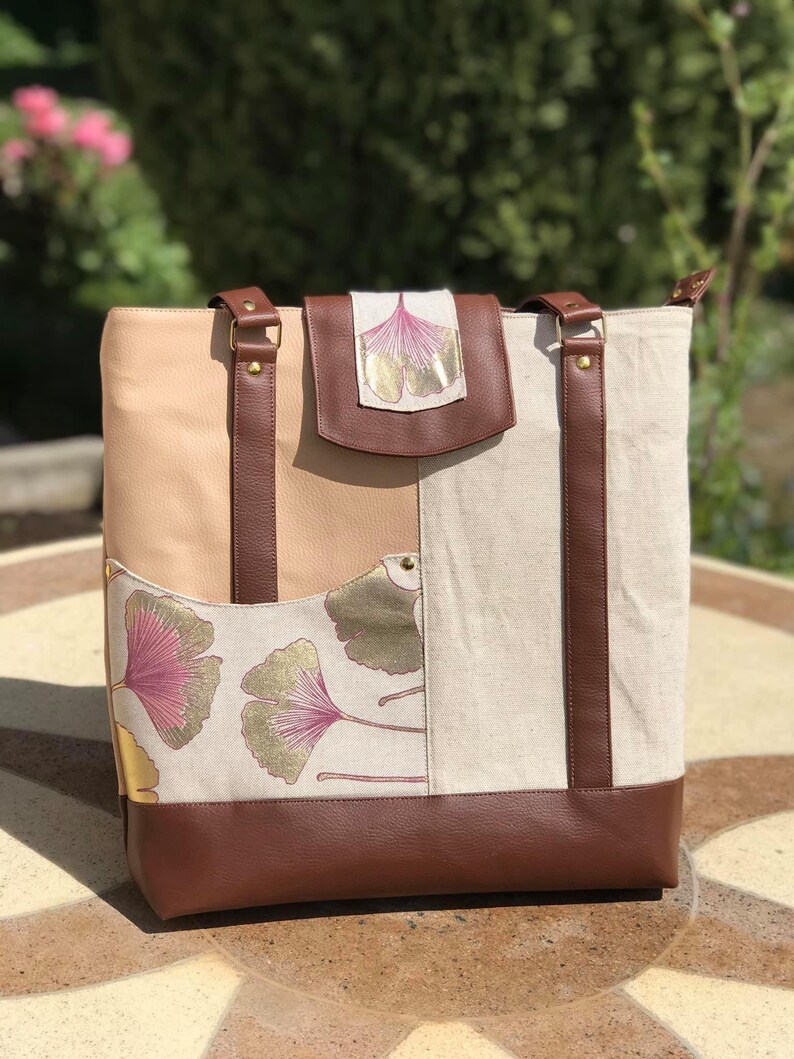 Puede incluir: Un bolso tote de cuero marr&oacute;n con un panel de tela beige y blanco con un dise&ntilde;o de hoja de ginkgo. El bolso tiene dos correas de cuero marr&oacute;n y una solapa de cuero marr&oacute;n con un dise&ntilde;o de hoja de ginkgo.
