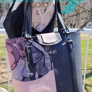 Puede incluir: Un bolso tote negro con estampado floral rosa y negro. El bolso tiene dos asas negras y un cierre de cremallera. El bolso tiene un peque&ntilde;o bolsillo rosa en la parte delantera y una etiqueta de cuero negro con el texto "Dreamscape Studio" en ella.