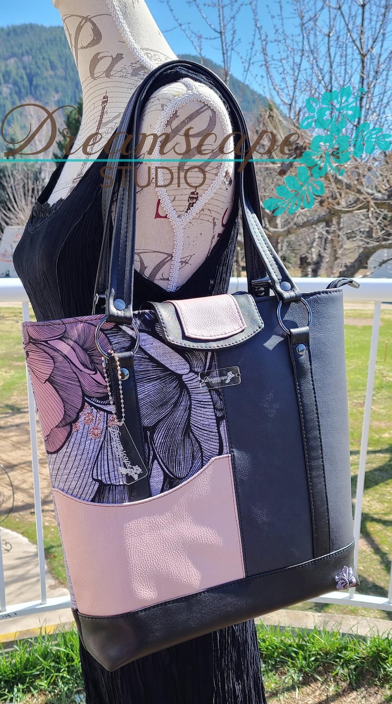 Puede incluir: Un bolso tote negro con estampado floral rosa y negro. El bolso tiene dos asas negras y un cierre de cremallera. El bolso tiene un peque&ntilde;o bolsillo rosa en la parte delantera y una etiqueta de cuero negro con el texto "Dreamscape Studio" en ella.