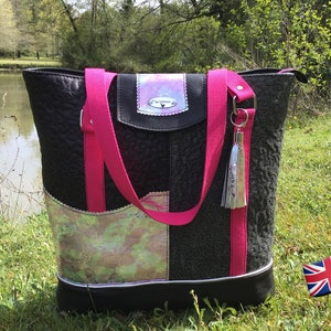 Puede incluir: Un bolso tote negro con correas rosas y un borla plateado. El bolso tiene un dise&ntilde;o de patchwork con un panel de tela iridiscente y una solapa de cuero negro con un logotipo plateado.
