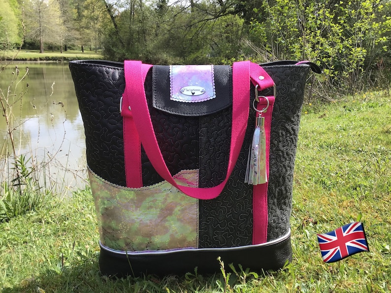 Puede incluir: Un bolso tote negro con correas rosas y un borla plateado. El bolso tiene un dise&ntilde;o de patchwork con un panel de tela iridiscente y una solapa de cuero negro con un logotipo plateado.