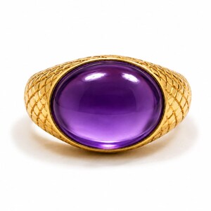 May include: A gold ring with a large, oval, purple gemstone. The ring's band has a textured, diamond-patterned design. The gemstone is a vibrant shade of purple, set in a gold bezel. The ring is displayed on a white background.