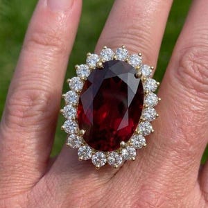 May include: A gold ring featuring a large, oval-cut deep red gemstone at the center, surrounded by a halo of twenty-two small, round, clear white stones. The setting is crafted from yellow gold, highlighting the contrast between the dark red center.