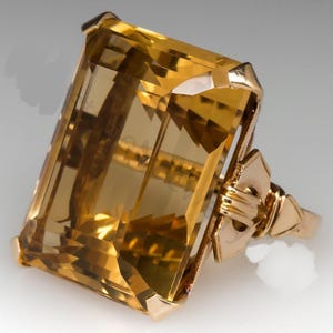 Könnte beinhalten: Ein goldener Ring mit einem großen, rechteckig geschliffenen Citrin-Edelstein. Der Citrin hat eine warme, goldgelbe Farbe und ist in einem Goldband mit aufwendigen Details gefasst. Der Ring hat ein klassisches, elegantes Design.