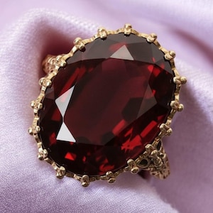 Puede incluir: Un anillo dorado con una gran piedra preciosa roja oscura de talla ovalada y facetada. El engaste del anillo tiene un diseño ornamentado y texturizado con pequeños elementos decorativos. El anillo se muestra sobre una tela lavanda suave.