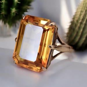 Puede incluir: Un anillo de oro con una gran piedra preciosa rectangular amarilla. La piedra preciosa tiene un corte facetado y está engastada en un engaste simple y elegante.