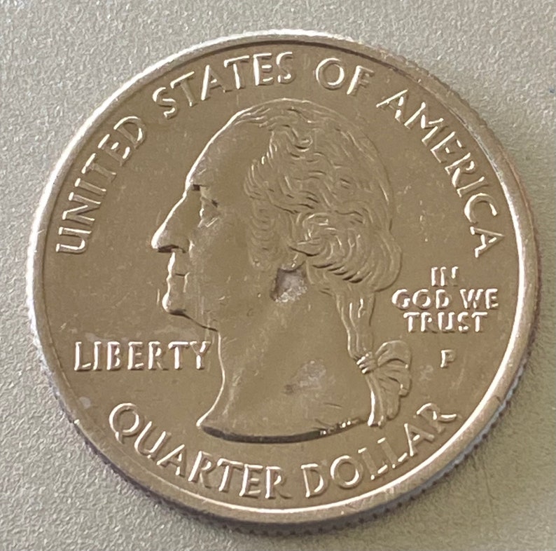 Puede incluir: Una moneda de veinticinco centavos de d&oacute;lar estadounidense. El anverso presenta un perfil de George Washington, las inscripciones "LIBERTY", "QUARTER DOLLAR", "UNITED STATES OF AMERICA", y el lema "IN GOD WE TRUST".