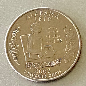 May include: A United States quarter coin featuring Helen Keller seated, with the text "Alabama 1819" at the top. The coin also includes the words "Spirit of Courage" and the year 2003.