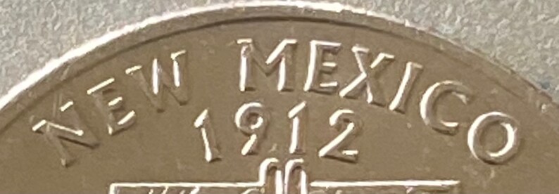 Puede incluir: Primer plano de una moneda de plata con la inscripci&oacute;n "NEW MEXICO 1912". El texto est&aacute; en relieve sobre la superficie de la moneda. El dise&ntilde;o de la moneda presenta una vista parcial del nombre del estado y el a&ntilde;o de su admisi&oacute;n.