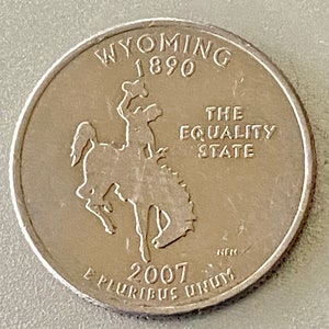 May include: A silver quarter coin featuring a bucking bronco and rider. The coin is from Wyoming, with the text "Wyoming 1890" at the top and "The Equality State" on the right. The year 2007 is at the bottom.