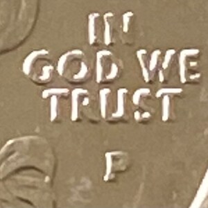 Puede incluir: Primer plano de una moneda dorada con la inscripci&oacute;n "IN GOD WE TRUST" en may&uacute;sculas. La letra "P" es visible debajo del texto. La superficie de la moneda tiene un aspecto ligeramente desgastado.