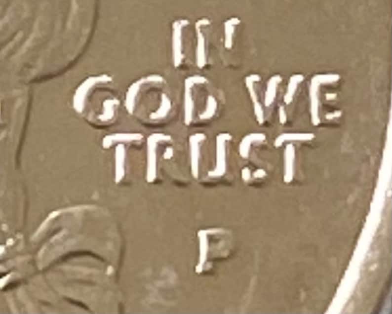 Puede incluir: Primer plano de una moneda dorada con la inscripci&oacute;n "IN GOD WE TRUST" en may&uacute;sculas. La letra "P" es visible debajo del texto. La superficie de la moneda tiene un aspecto ligeramente desgastado.