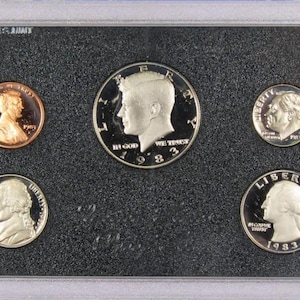 May include: A 1983 United States proof set of coins, including a copper penny, a silver half dollar, a dime, a nickel, and a quarter. The coins are displayed in a protective case with a black background.
