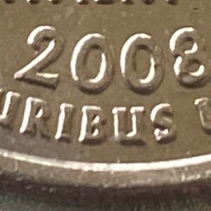 Puede incluir: Primer plano de una moneda plateada con el a&ntilde;o 2008 visible. La inscripci&oacute;n "E PLURIBUS UNUM" es visible en el borde. La superficie de la moneda muestra un ligero desgaste.