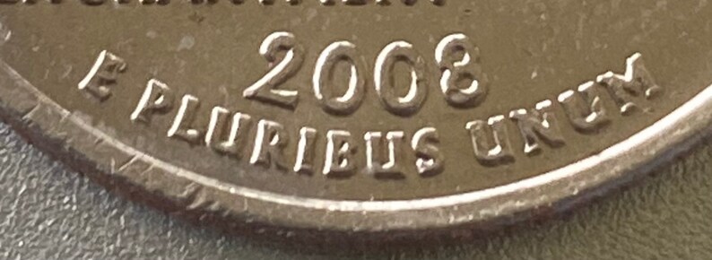 Puede incluir: Primer plano de una moneda plateada con el a&ntilde;o 2008 visible. La inscripci&oacute;n "E PLURIBUS UNUM" es visible en el borde. La superficie de la moneda muestra un ligero desgaste.