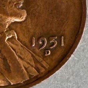 May include: A close-up view of a copper-colored United States one-cent coin showing the year 1951 and a D mint mark located directly below the date. The surface of the coin displays a metallic texture with visible raised relief details of a figure.