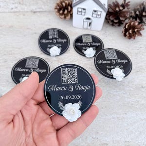 May include: A circular black acrylic disc featuring a white QR code, the text "Marco & Ronja", the date "26.09.2026", and a small white artificial flower attached to the bottom. The surface is smooth and reflective with a thin white border.