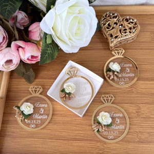 May include: Gold-colored ring-shaped wedding favors with a diamond-shaped top, featuring the names "Exora & Martin" and the date "09.13.25". Each ring is decorated with a white rose and floral accents. A heart-shaped box is also visible.