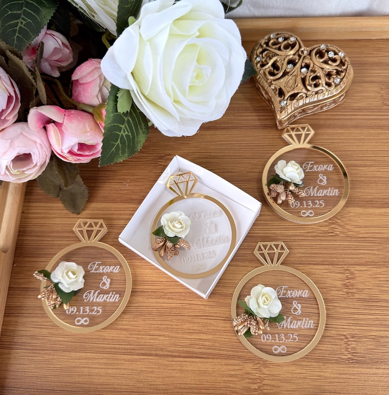 May include: Gold-colored ring-shaped wedding favors with a diamond-shaped top, featuring the names "Exora & Martin" and the date "09.13.25". Each ring is decorated with a white rose and floral accents. A heart-shaped box is also visible.