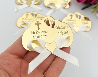 May include: Gold-colored acrylic cloud-shaped favor with a white ribbon and a small gold heart. The cloud has a cross, two footprints, and the text "Mi Bautizo 10.07.2023 Brianna Ayala".