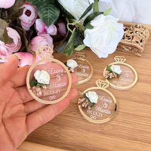 May include: Clear acrylic wedding favors shaped like rings with gold accents. Each ring features a white rose, gold floral details, and the names "Exora & Martin" with the date "09.13.25". The infinity symbol is also included.